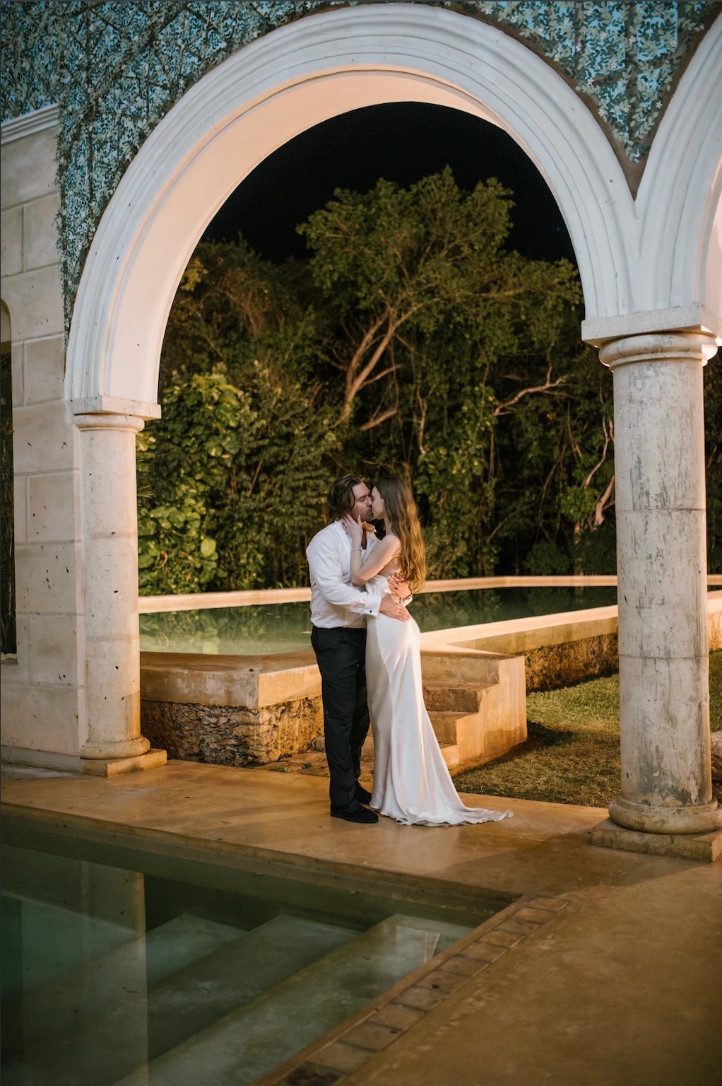 Amy and John kissing beneath a stone arch by a pool at night.